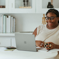 A smiling Black female worker, dressed in a white outfit and holding a coffee mug with both hands, while working from home