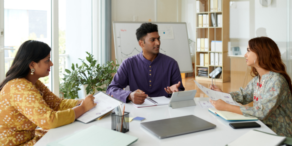 Three Indians sitting at a table, discussing documents and using a tablet in a modern office setting