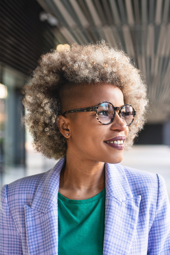 A woman with blonde curly hair and glasses smiles as she looks to the side. She is wearing a purple checkered blazer over a green shirt and stands outdoors in front of a modern building.